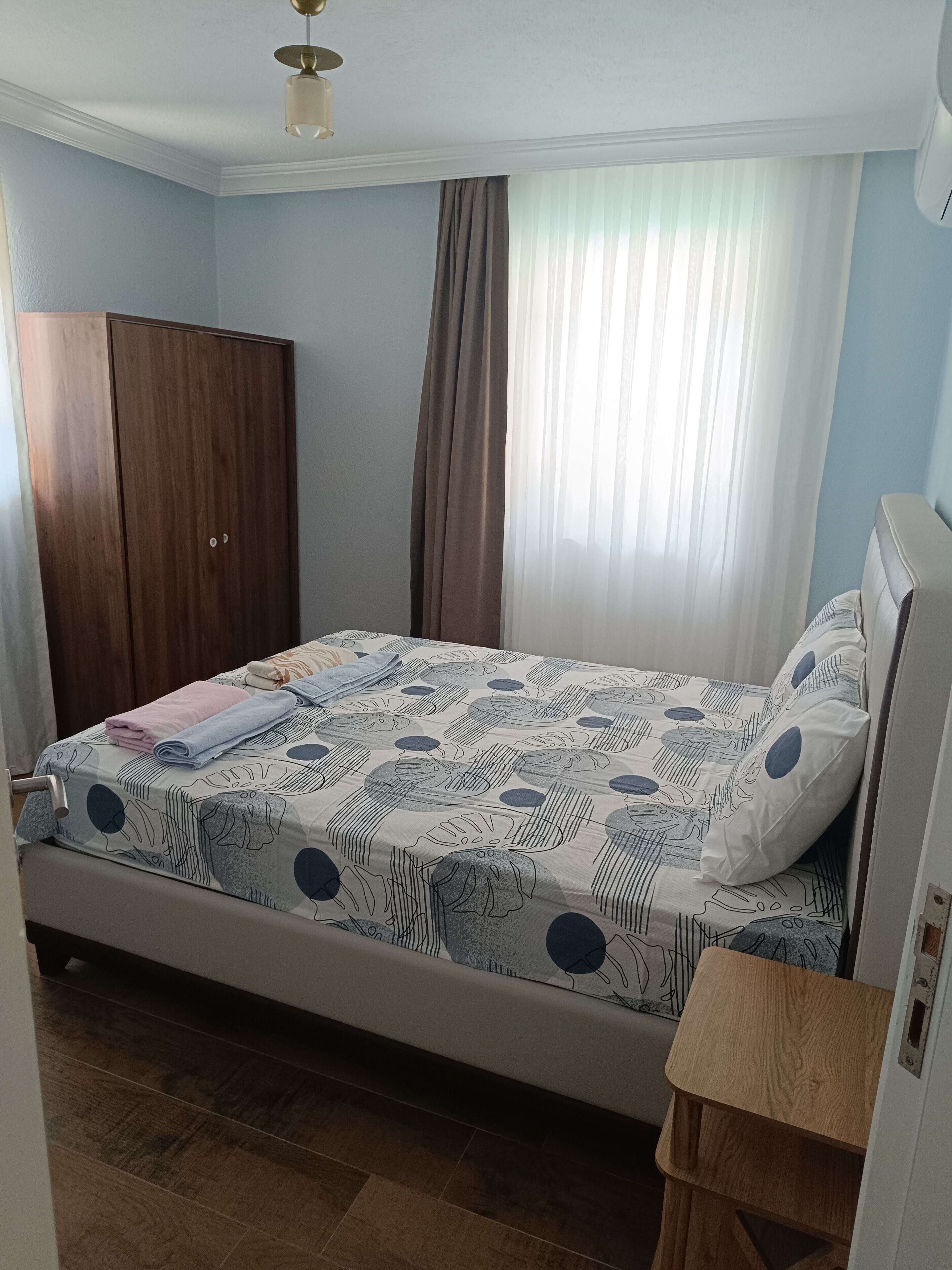 A bedroom with a bed, a wooden wardrobe, a bedside table, and a window with curtains.