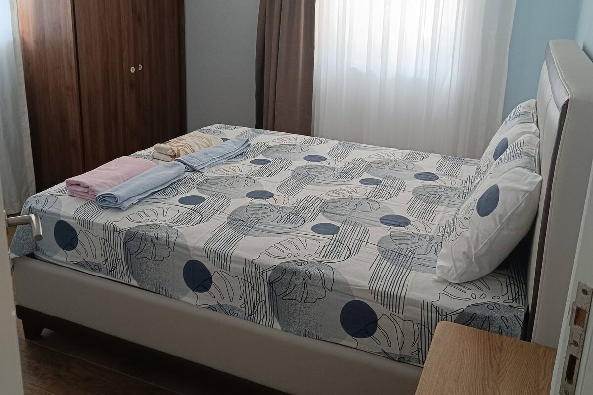 A bedroom with a bed, a wooden wardrobe, a bedside table, and a window with curtains.