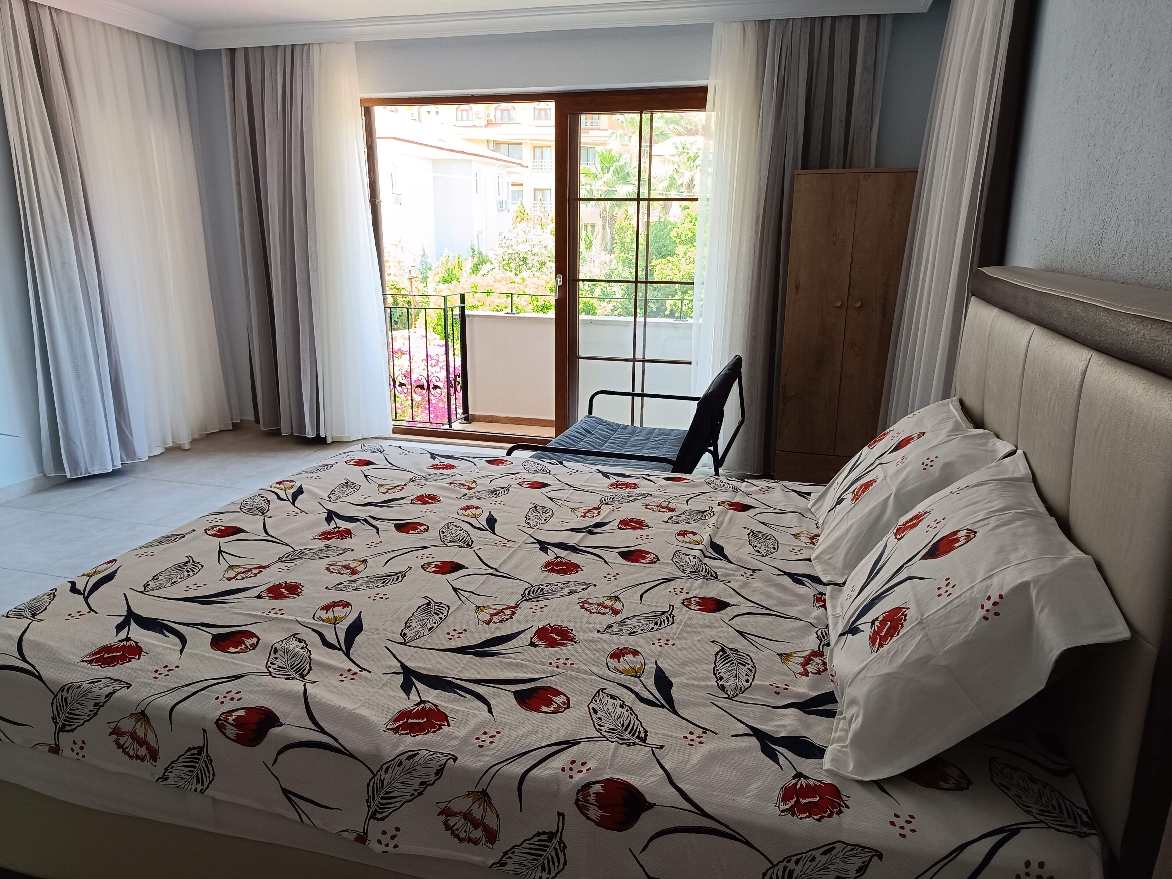 A hotel room with a bed, a chair, a balcony with a view of buildings and greenery.