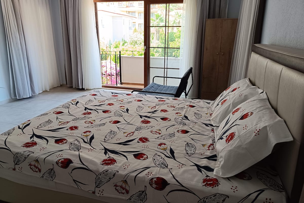 A hotel room with a bed, a chair, a balcony with a view of buildings and greenery.