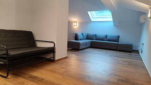 Quarto no estilo loft com piso de madeira, estrutura de cama em metal preto, sofá seccional cinza e um claraboia.