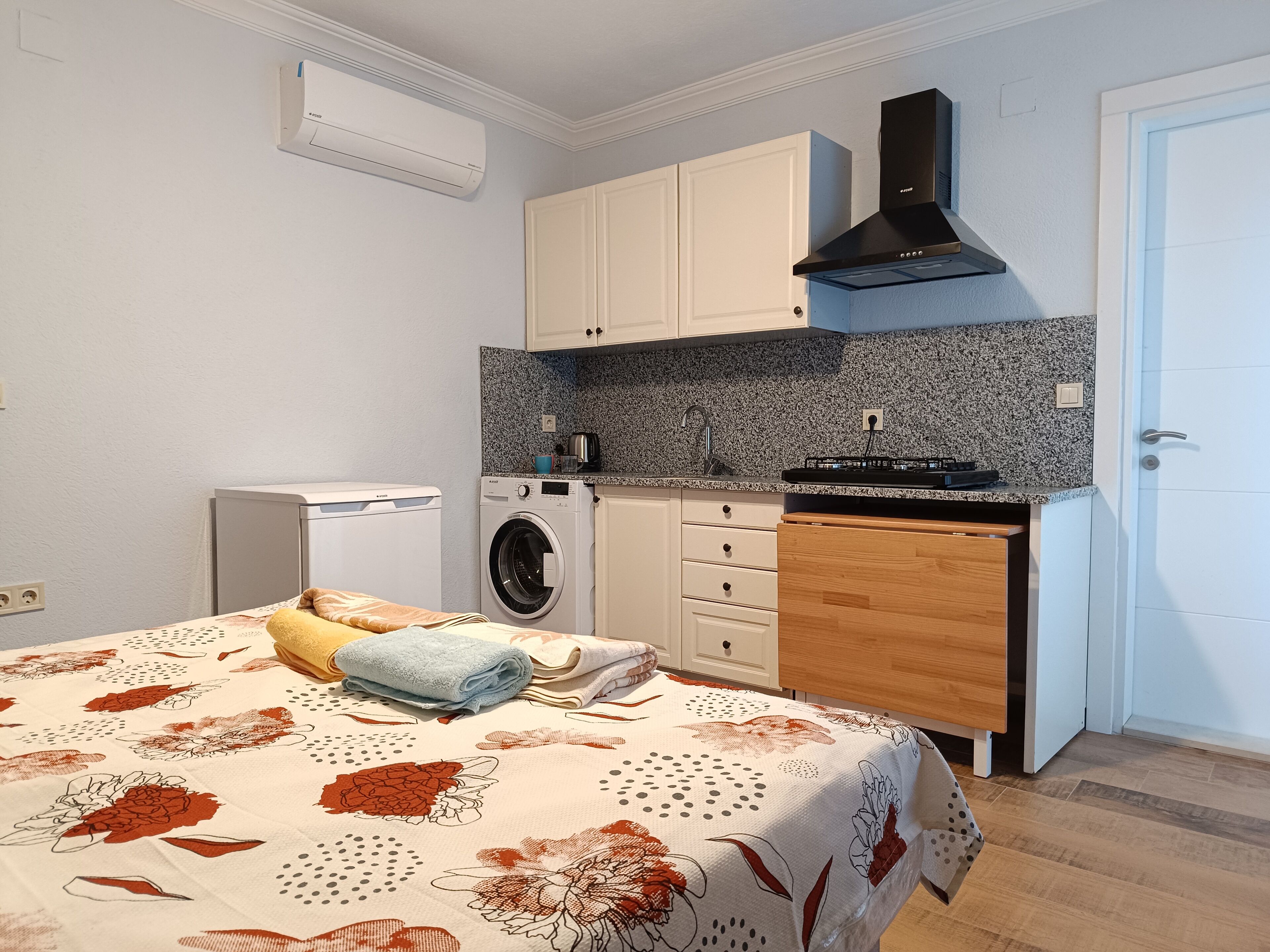 A compact room with a bed, a washing machine, a small kitchen with a stove, and a door.