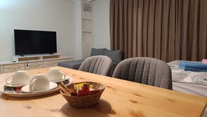 Quarto de hotel com mesa de madeira, duas poltronas cinzentas, um sofá, uma TV e uma janela com cortinas.