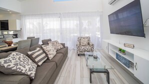Una sala moderna con un sofá, un sillón, una mesa de centro de vidrio y un televisor montado en la pared.