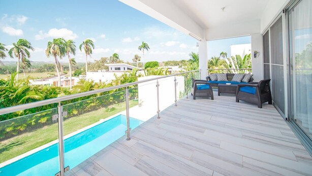 Un balcón con piscina, mobiliario exterior y vistas a palmeras y edificios.