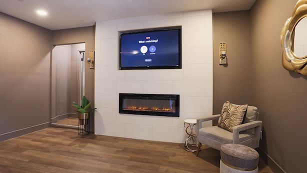 Una sala moderna con una televisión de pantalla plana sobre la chimenea, un área de descanso confortable con sofá y reposapiés, y una planta en maceta.