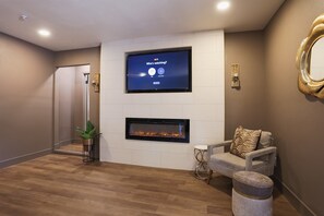 A modern living room with a flat-screen TV mounted above a fireplace, a comfortable seating area with a sofa and ottoman, and a potted plant.