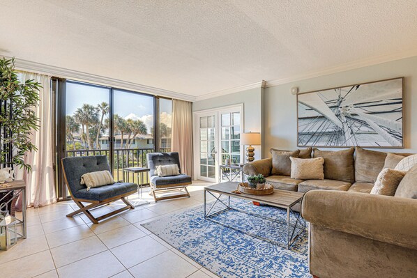 A spacious living room with a large window, a sofa, a chair, and a coffee table. - Luxe Fort Pierce Beach Condo w/ Golf Views! (Fort Pierce)