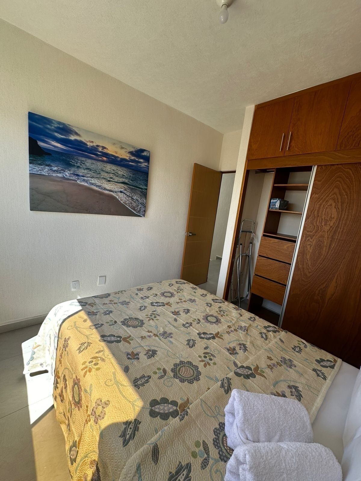 A bedroom with a bed, a wooden wardrobe, a painting of a beach, and a small cabinet.