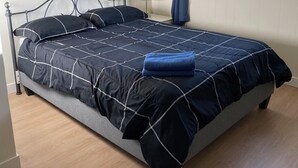 Een net opgemaakt bed met een donkerblauwe en zwarte quilt, een grijze kussen en een blauwe handdoek erop. Er zijn twee bloemschilderijen aan de muur en een raam met blauwe gordijnen.