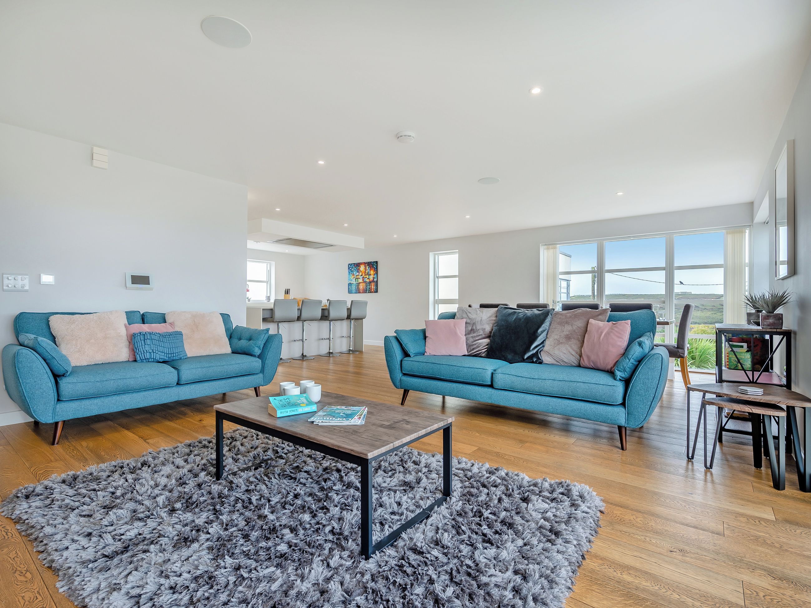 A spacious living room with two blue sofas, a coffee table, and a view of the kitchen and outdoor scenery.