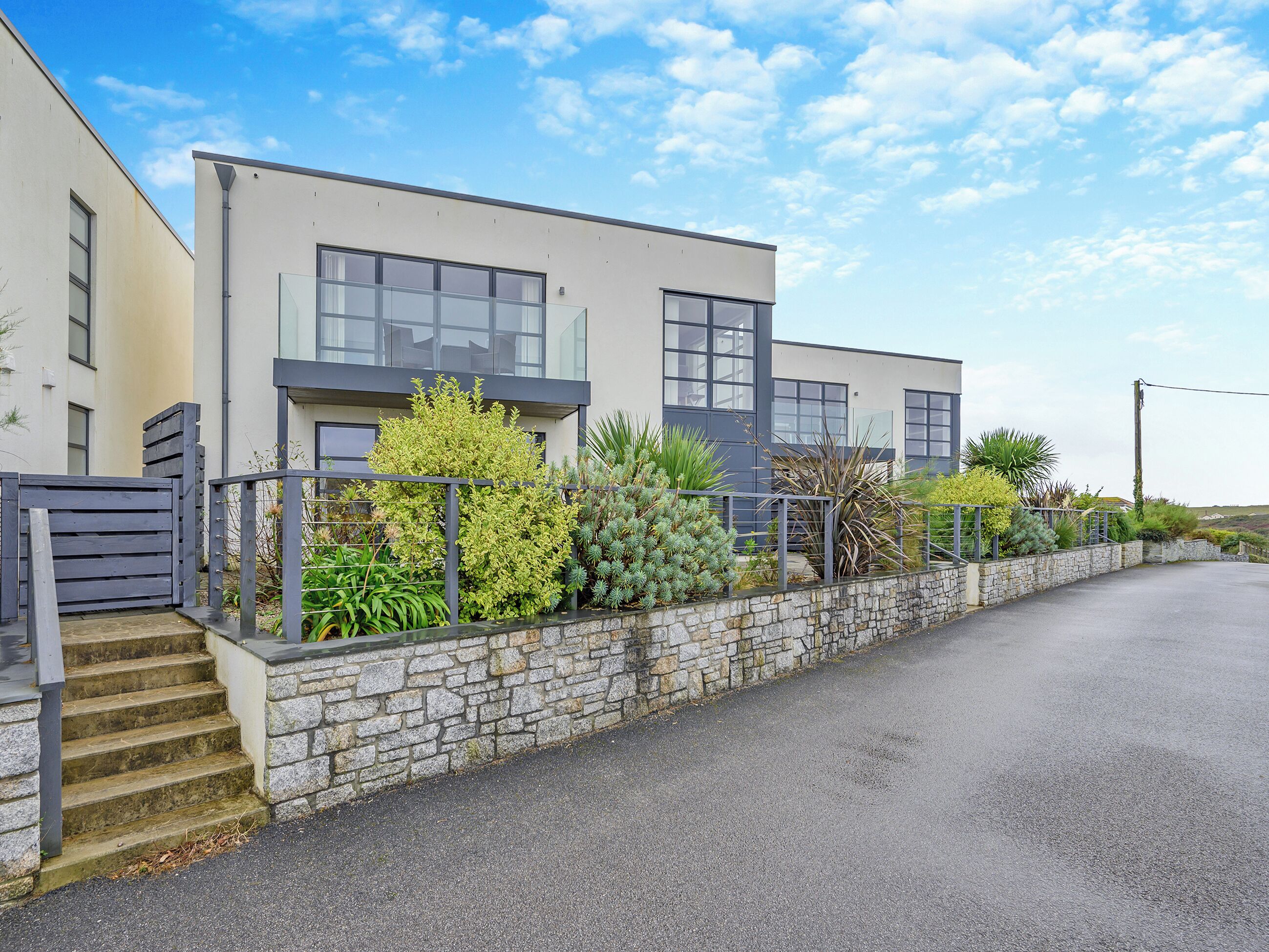 A modern two-story building with a stone wall and metal railing, featuring large windows and a variety of plants.