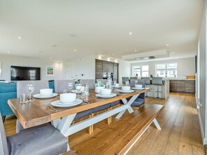 Un comedor moderno con una mesa de madera dispuesta para cuatro personas, sillas grises tapizadas y una cocina al fondo.