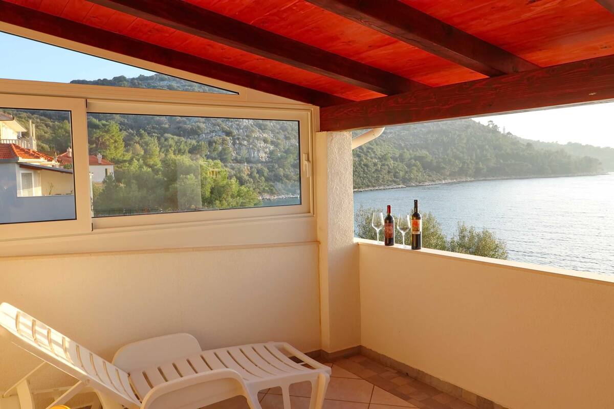 A balcony with a view of a lake and hills, a white chair, and two bottles of wine.