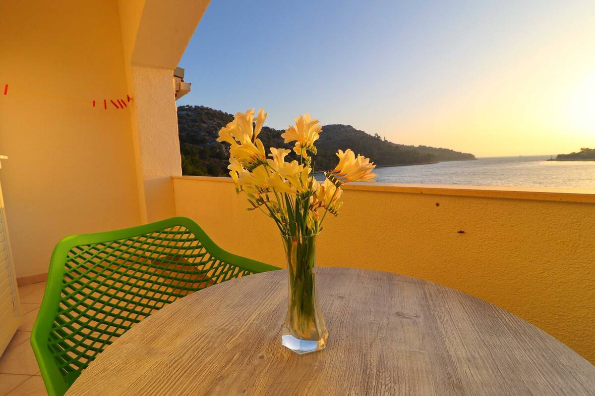 A balcony with a wooden table and a vase of yellow flowers, overlooking a body of water and a hilly landscape.