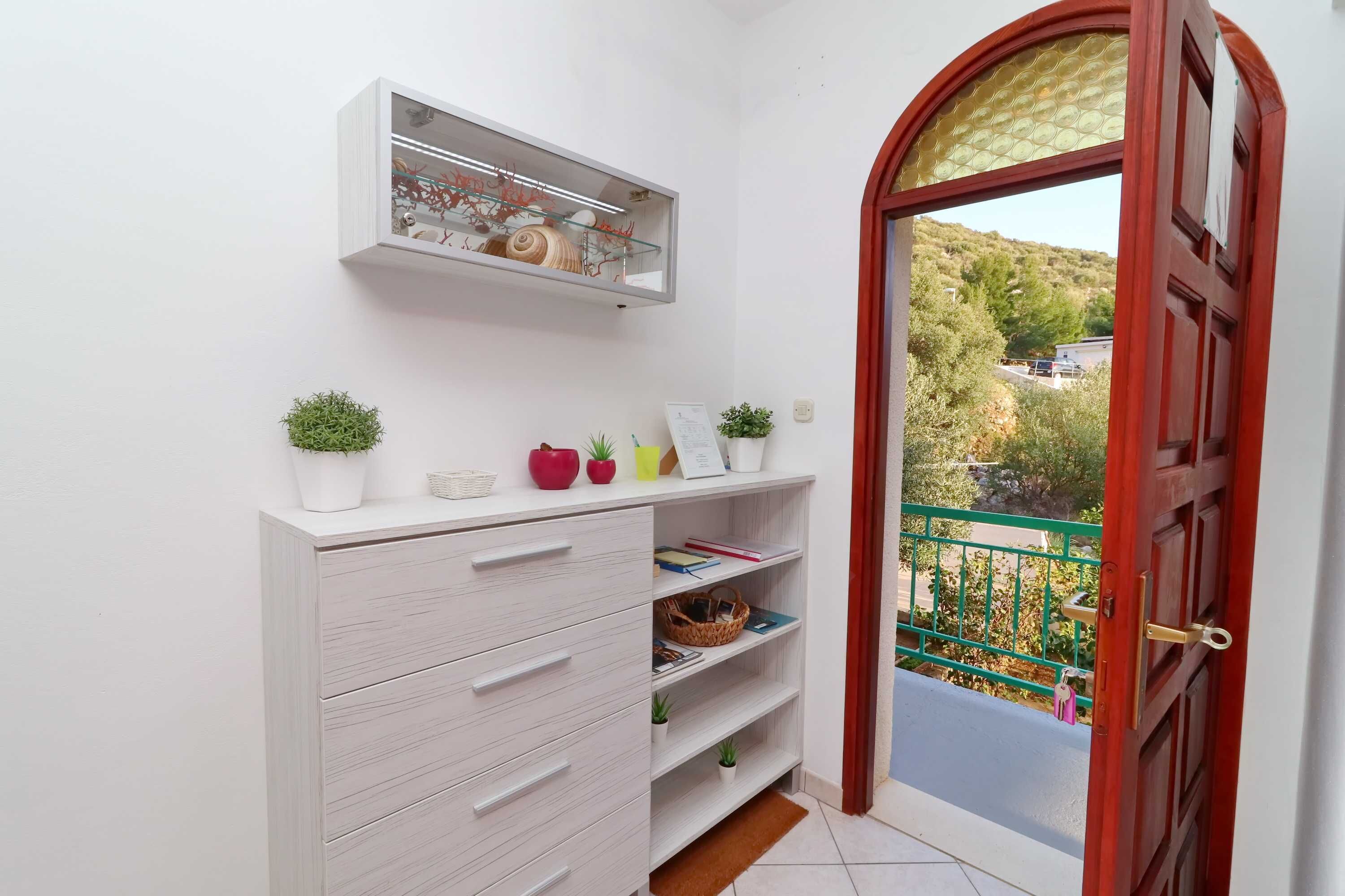 A white interior with a wooden door, a shelf with decorative items, and a view of greenery outside.