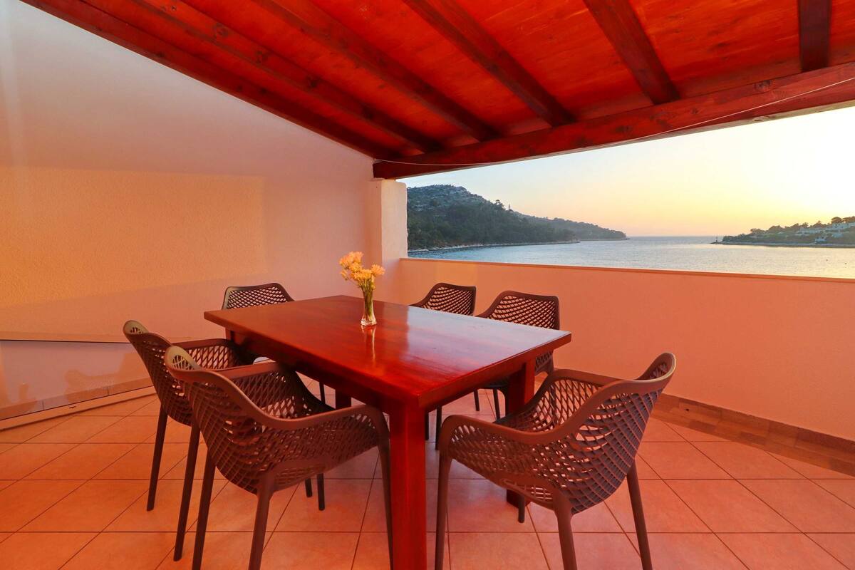 A dining area with a wooden table and wicker chairs, overlooking a body of water and a distant hill.