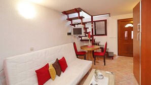 Una sala de estar con un sofá blanco, cojines rojos y marrones, una pequeña mesa de madera con sillas, un televisor y una puerta.