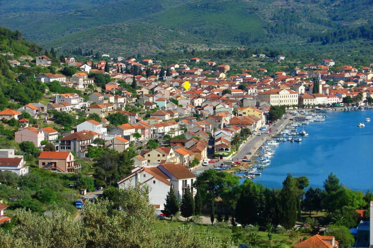 A coastal town with a marina, houses with red roofs, and green hills in the background.