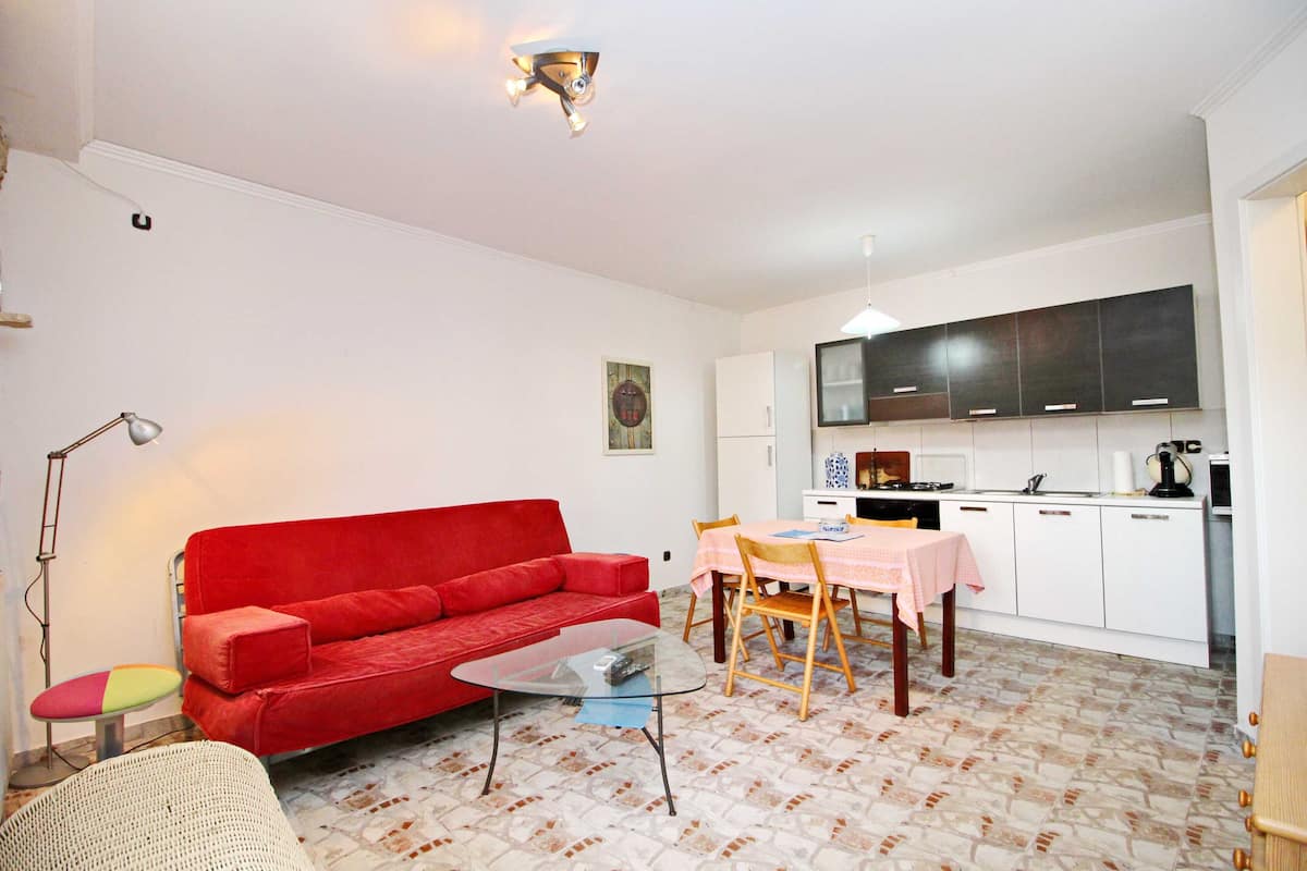 A compact living space with a red sofa, a small table, a dining table with chairs, a kitchen area, and a patterned floor.