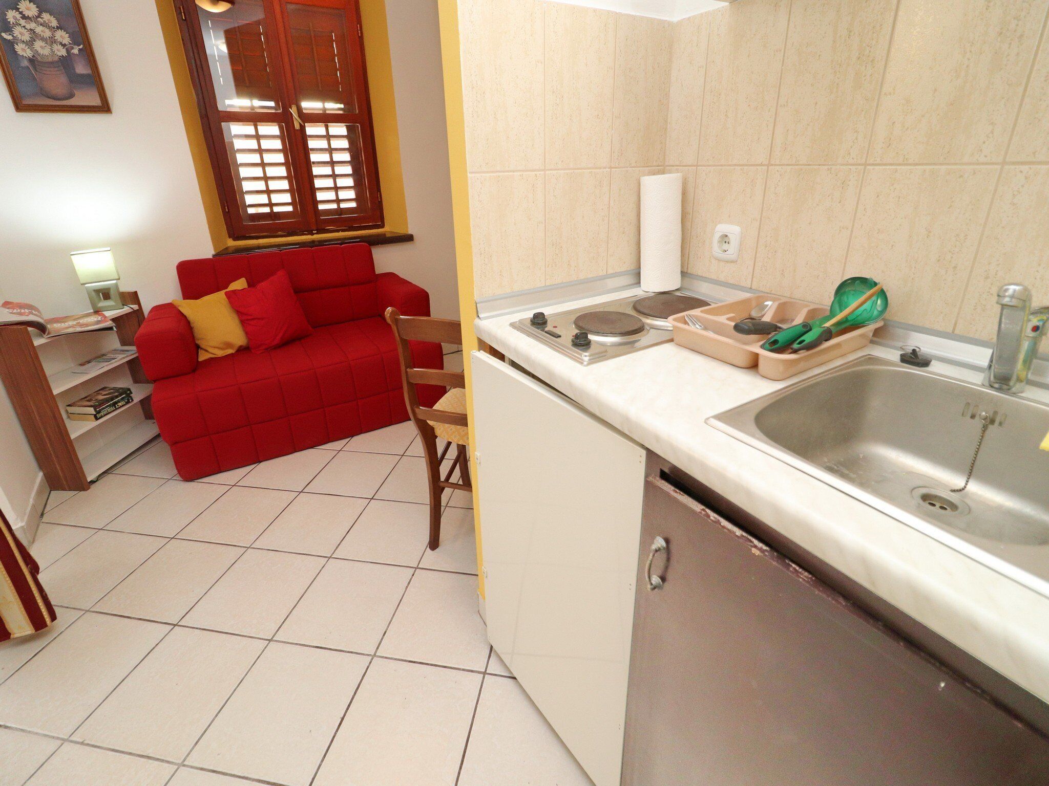 A compact kitchen with a sink, stove, and countertop. A red sofa and wooden chair are in the adjacent living area.