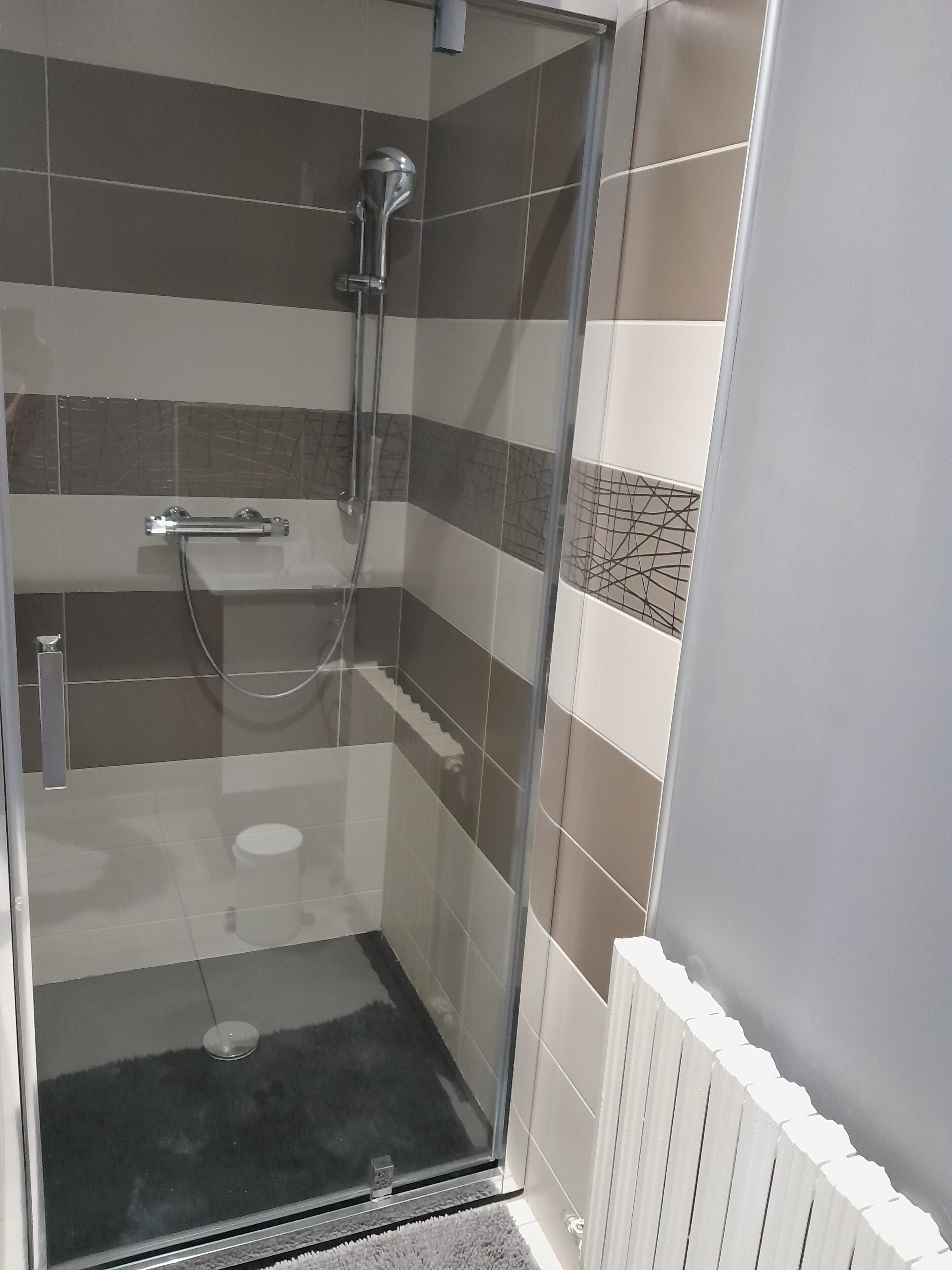 Una cabina doccia in vetro con soffione a mano, rubinetto a parete e un piccolo gradino che conduce a un pavimento piastrellato.