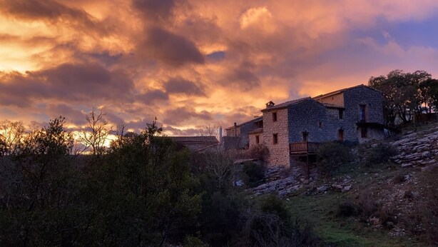 A stone building with a sloped roof, surrounded by vegetation and under a dramatic sunset sky. - Les Gîtes du mas Champion (Saint-Just-et-Vacquières)