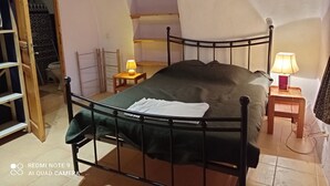A bedroom with a metal bed frame, a bedside table with a lamp, a wooden chair, and a small table with a lamp. There is a door leading to a bathroom with a toilet and a shower. - Les Gîtes du mas Champion (Saint-Just-et-Vacquières)