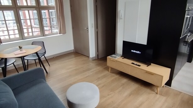 Una sala de estar moderna con un sofá gris, una mesita redonda, un televisor sobre un mueble de madera y un ventanal con cortinas.