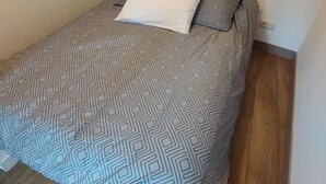 Una cama con una colcha de estampado geométrico y dos almohadas, una blanca y otra estampada, en una habitación con suelo de madera y pared lisa.