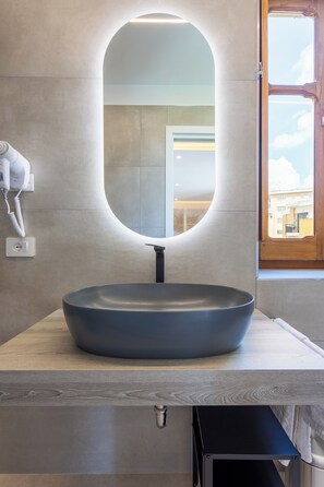 A modern bathroom with a large oval mirror, a round sink, and a hairdryer on the wall.