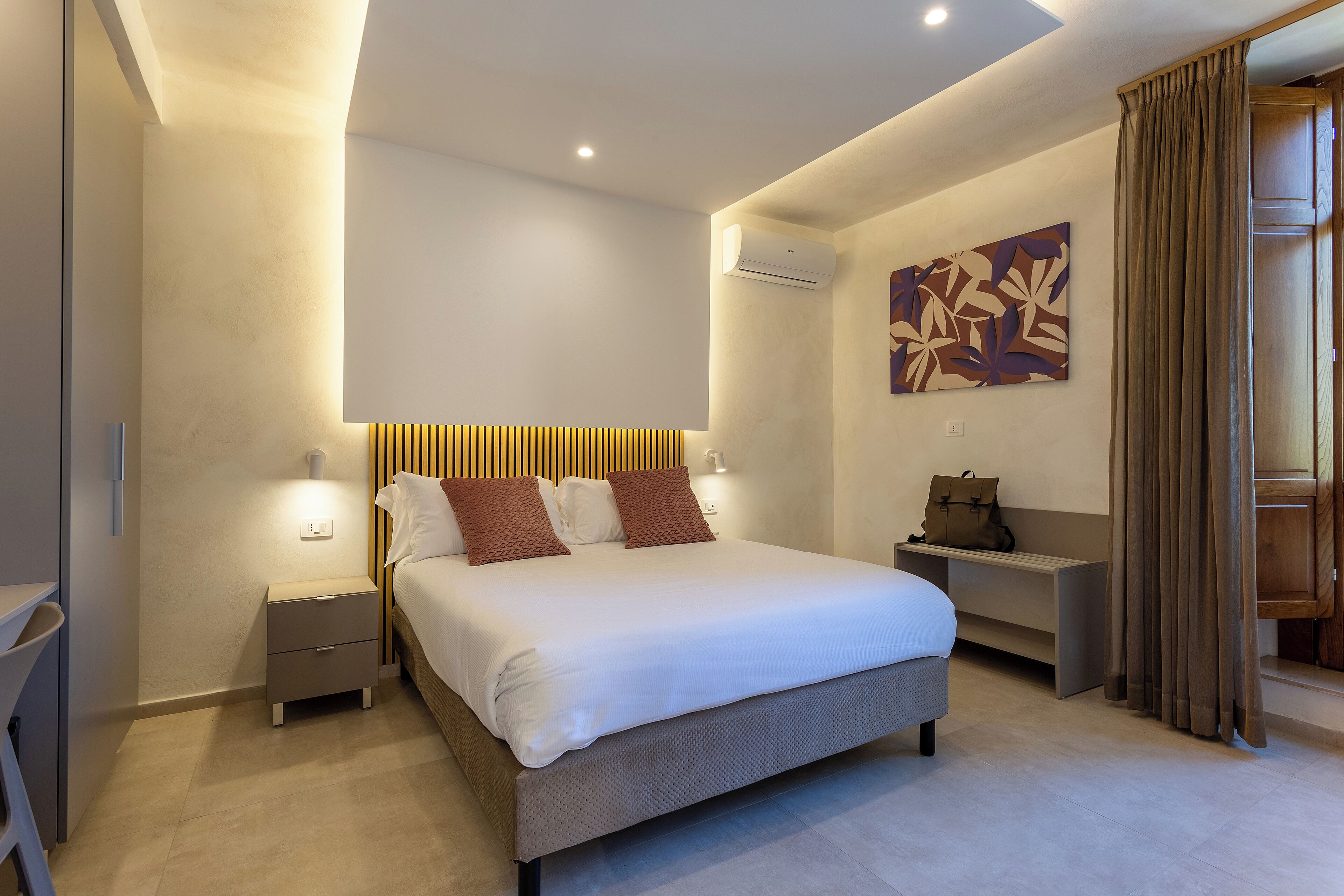 A modern hotel room with a large bed, bedside tables, a wall-mounted air conditioner, and a decorative wall piece.