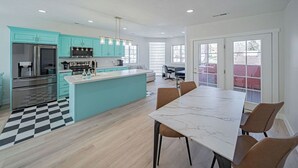 Een moderne keuken met blauwgroene kastjes, een marmeren eiland en een eethoek met een witte tafel en bruine stoelen.