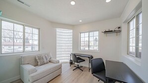 Een ruime kamer met een lichtgekleurde bank, een zwarte bureaustoel, een bureau en een groot raam met uitzicht op gebouwen.