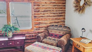 Una camera accogliente con una parete in legno, una poltrona in pelle marrone, un poggiapiedi decorato, un comodino in legno con una lampada e una pianta in vaso.