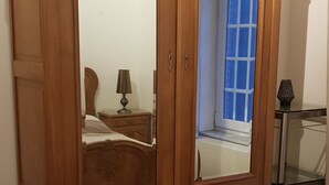 Une armoire en bois avec un miroir, une table de chevet avec une lampe et un lit recouvert d’une couverture blanche.