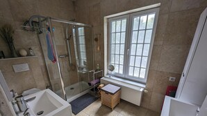 Une salle de bain moderne avec une douche à parois de verre, une fenêtre laissant entrer la lumière naturelle et un petit tabouret en bois.