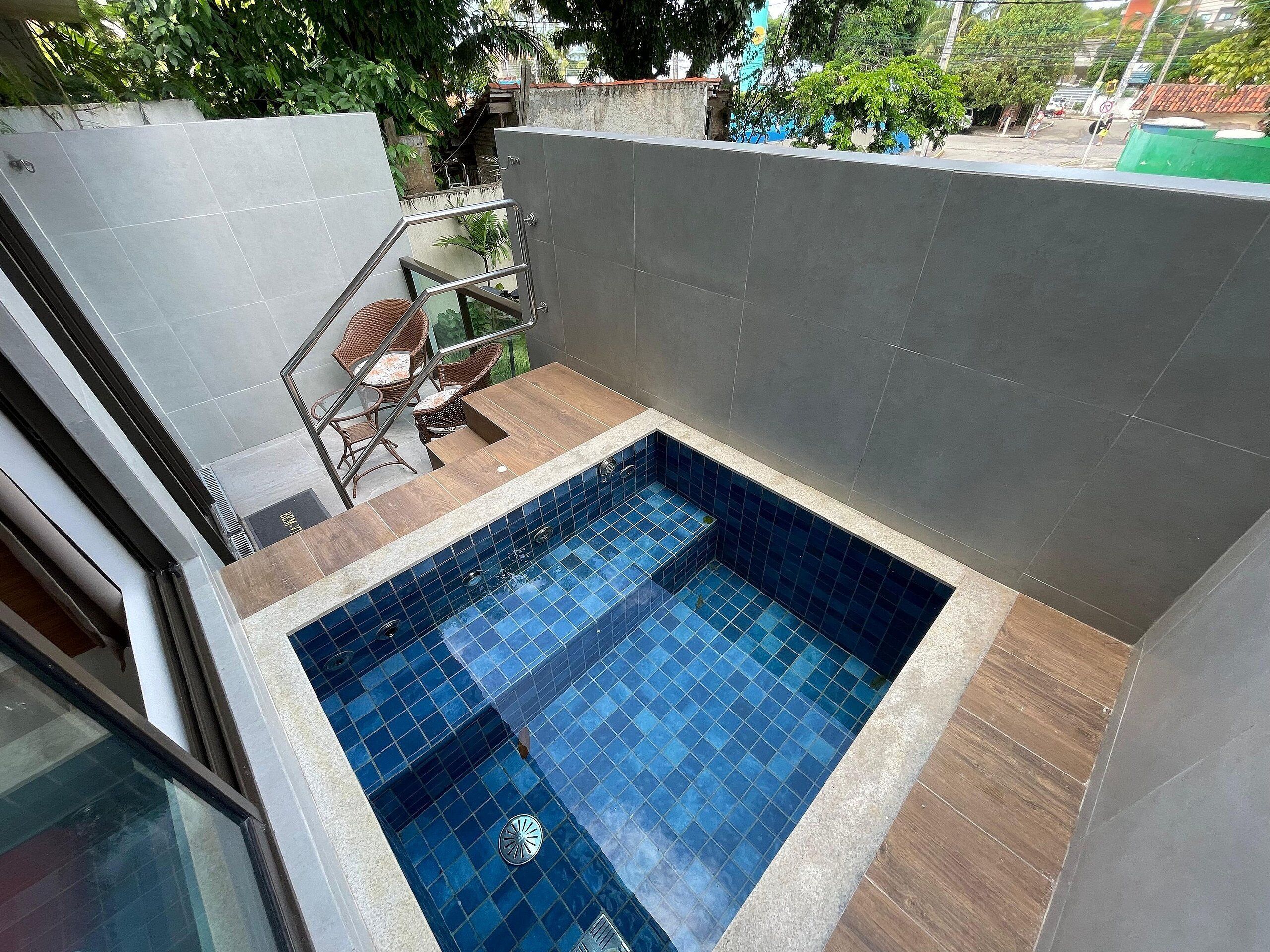 Une petite piscine carrée, aux carreaux bleus, et une zone de terrasse en bois avec des chaises.