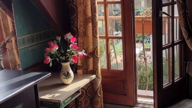 A corner of a room with a wooden desk, a vase with flowers, a patterned curtain, and a view of the outside through a glass door. - Belle Chambre Chaleureuse au Calme (Chennevières-sur-Marne)