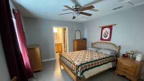 Un dormitorio con cama, cómoda de madera, mesita de noche, silla, ventilador de techo y puerta que da a otra habitación.