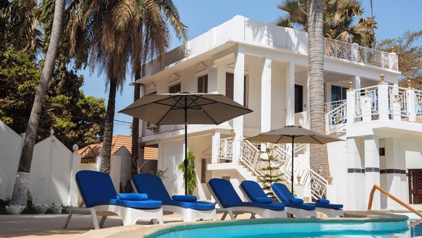 A pool area with lounge chairs and umbrellas, a white building with balconies, and palm trees. - Coconut Residence & Spa (Serrekunda)