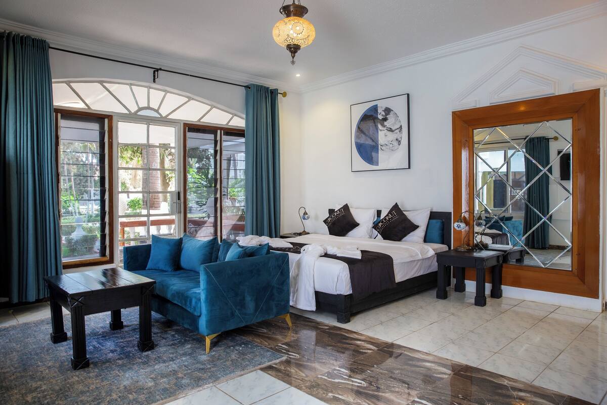 Quarto de hotel moderno com uma cama grande, um sofá azul, uma mesa pequena e uma janela ampla com cortinas.