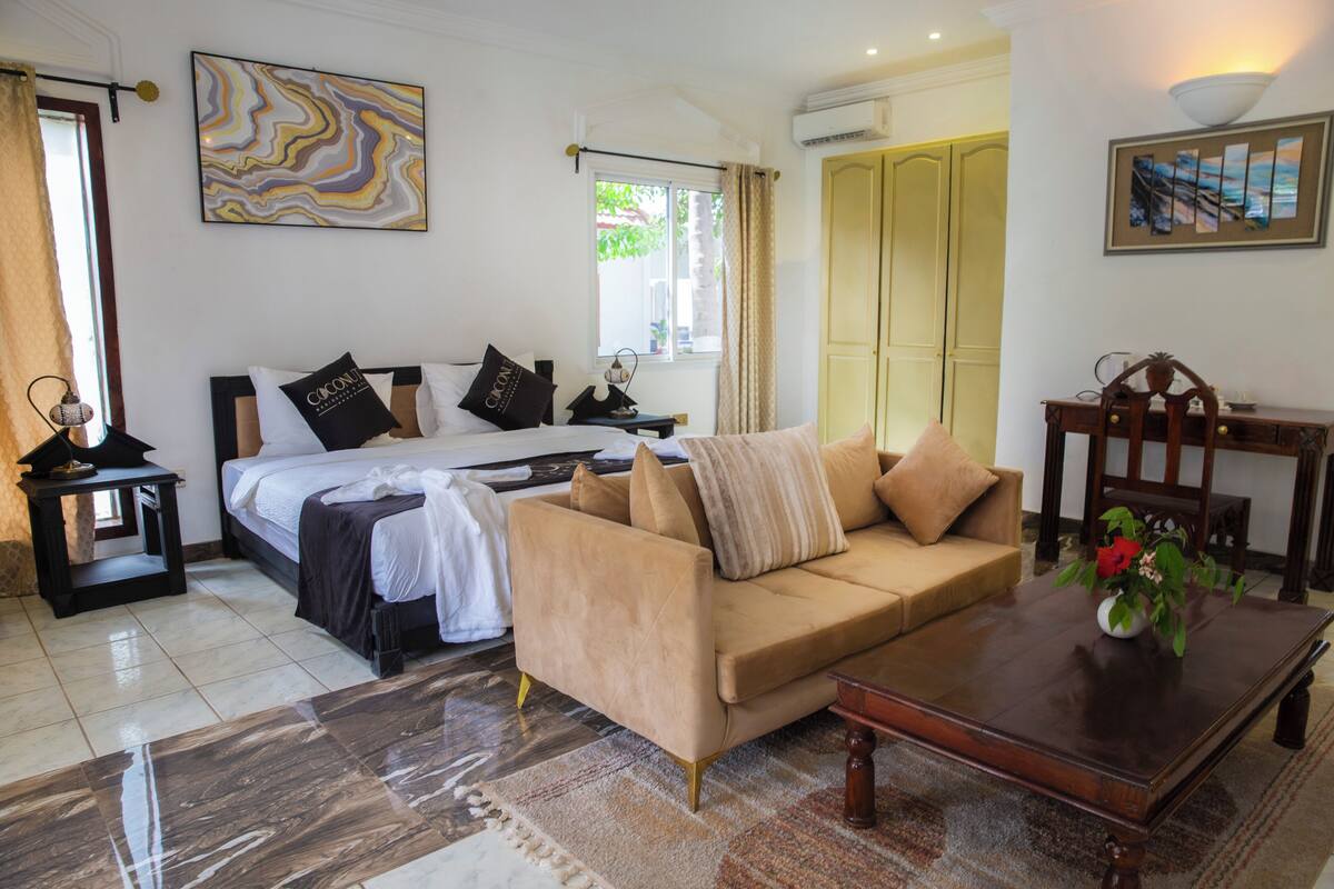 Quarto bem iluminado com uma cama, um sofá bege, uma mesa de centro e obras de arte nas paredes.