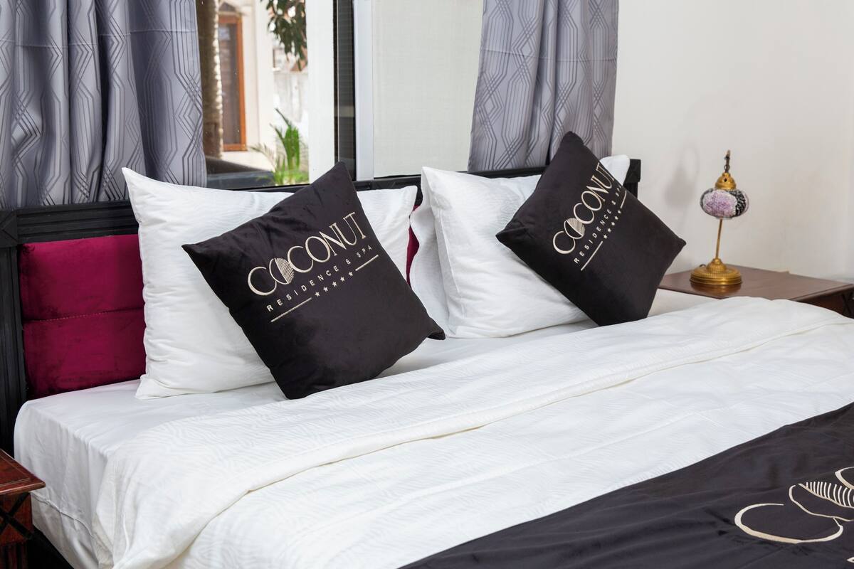 Cama bem arrumada com roupa de cama branca e duas almofadas pretas com o texto 'Coconuts Residences & Spa'.