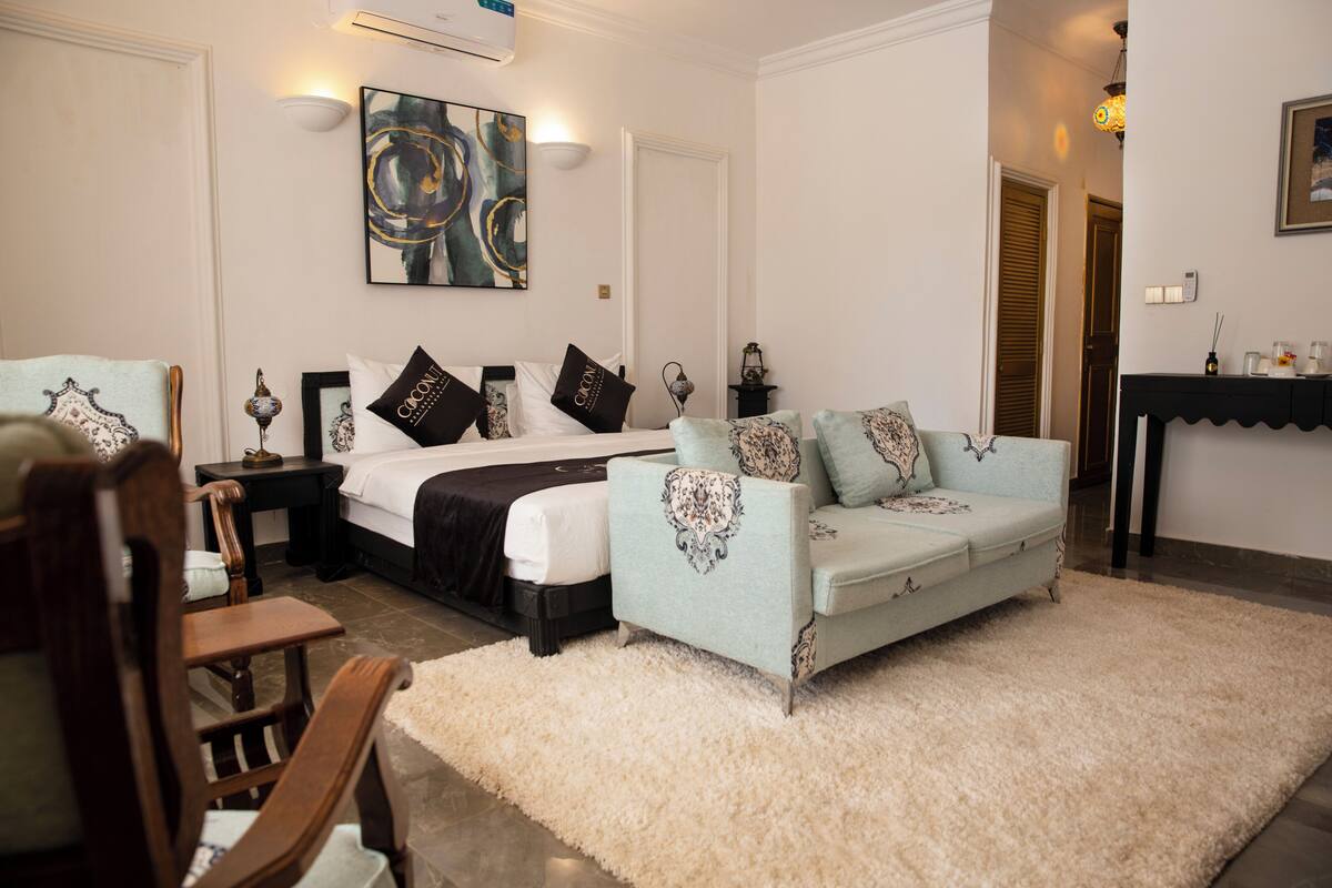 Quarto de hotel com uma cama, um sofá azul claro, uma cadeira de madeira, um tapete branco e um quadro na parede.