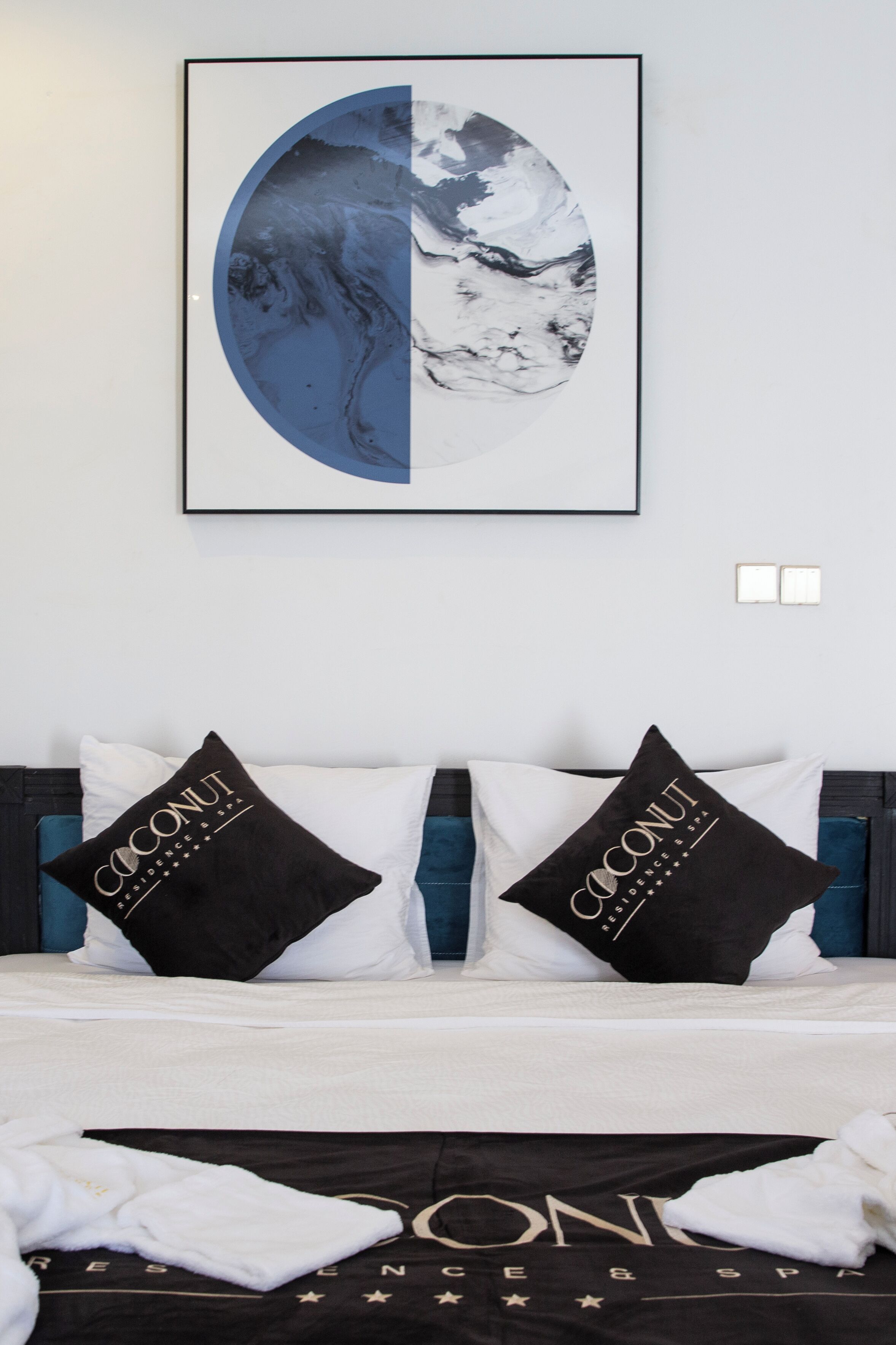 Una habitación de hotel con una cama, dos almohadas negras con la marca 'Coconut', un edredón blanco y una obra de arte enmarcada sobre la cama.