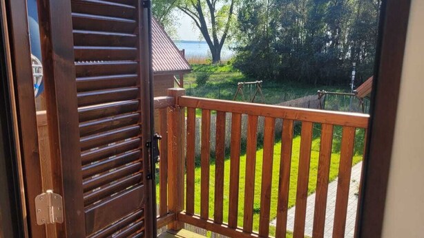 A wooden balcony with a checkered floor, overlooking a grassy area with trees and a body of water. - MoreView Kopice directly on the Szczecin Lagoon (Stepnica)