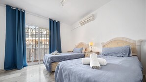 Habitación de hotel con dos camas, un balcón con cortinas azules, un aire acondicionado blanco y un cabecero de madera.
