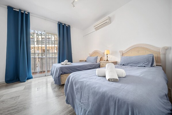 A hotel room with two beds, a balcony with blue curtains, a white air conditioner, and a wooden headboard.