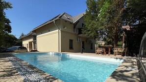 A swimming pool with a tiled deck, a house with a sloped roof, and a pergola with chairs. - See Haus - Podmaniczky (Balatonfoldvar)